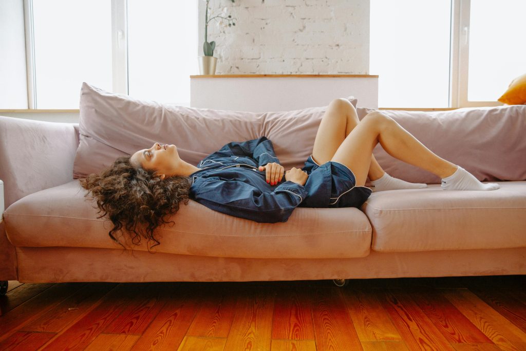 A woman in casual wear experiences abdominal discomfort while lying on a pink couch indoors.